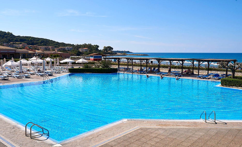 Blue outdoor swimming pool with sun loungers, parasols and the sea beyond at Aldemar Olympian Village.