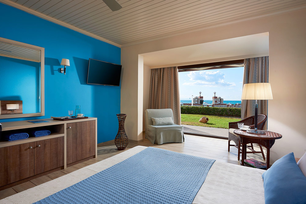 Double bungalow bedroom with blue accents and an open terrace facing a lawn and the sea at Aldemar Olympian Village.
